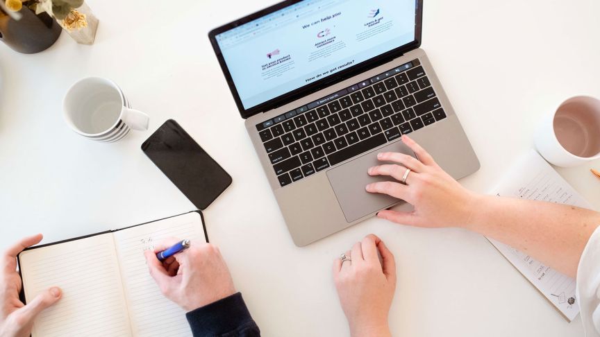 Photo of hands working on a notebook computer while someone else writes in a notebook