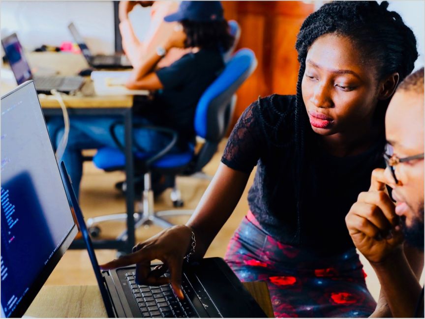 A person points at the screen of a notebook computer while another person looks on